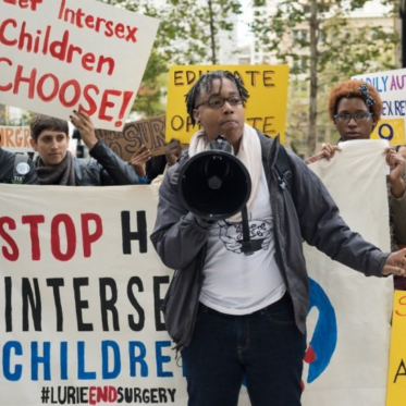 A person with a megaphone standing in front of a group of protesters who are holding signs with messages supporting intersex children's rights.